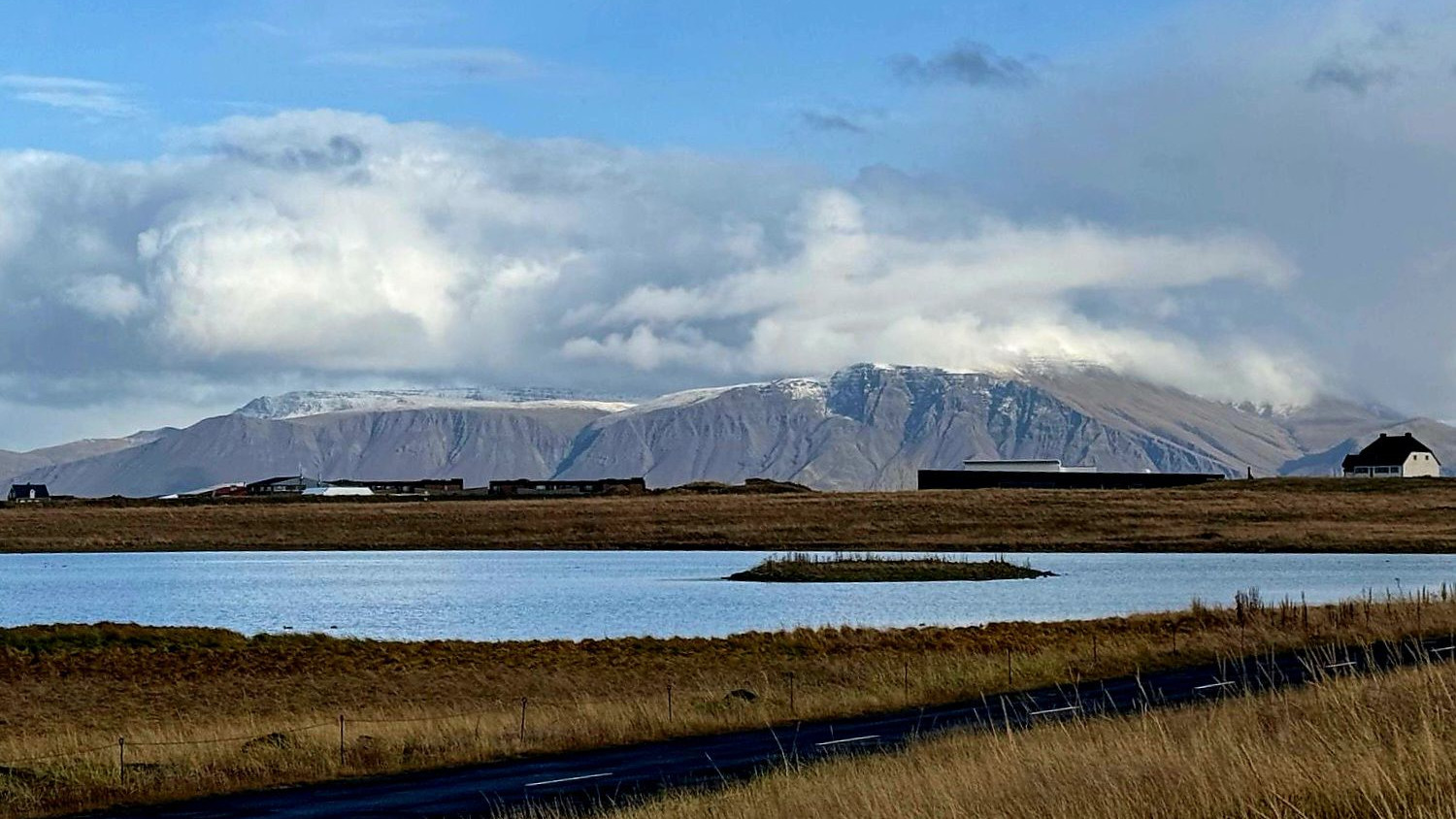 Mountains in the distance with a sky cluttered with clouds but blue overhead. In the foreground is marshland with a body of water. The grass that can be seen is brown. Taken from the Seltjarnarnes Peninsula in Iceland.