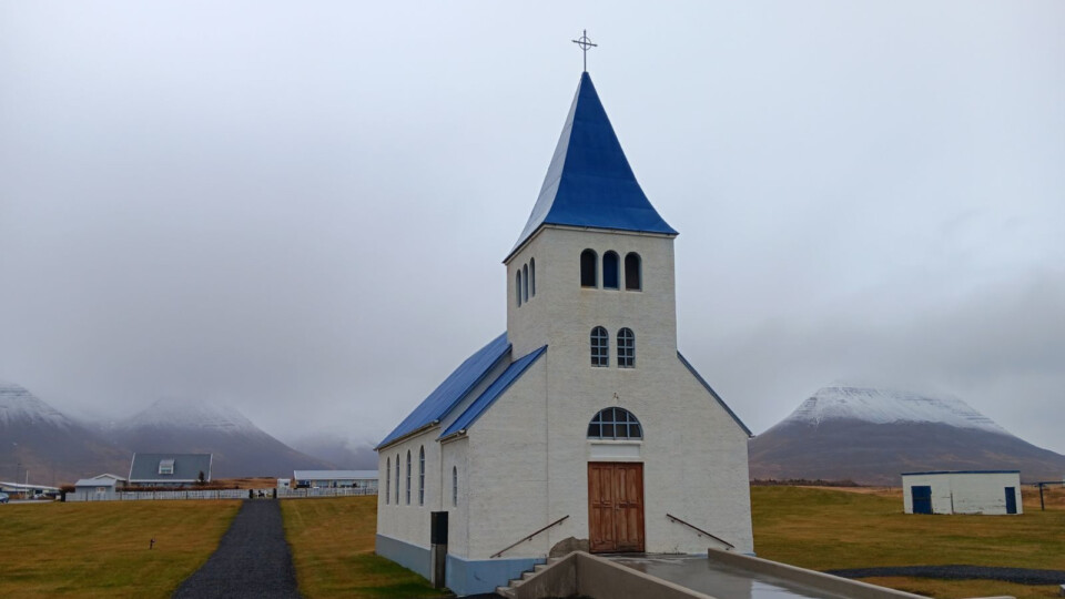 A small white church, with a blue roof. Mountainous, fog covered hills in the background.