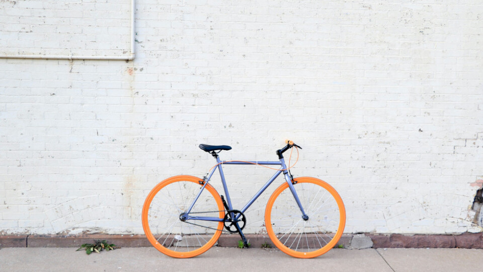 A blue framed single speed bicycle with orange wheels, against a brick wall painted white