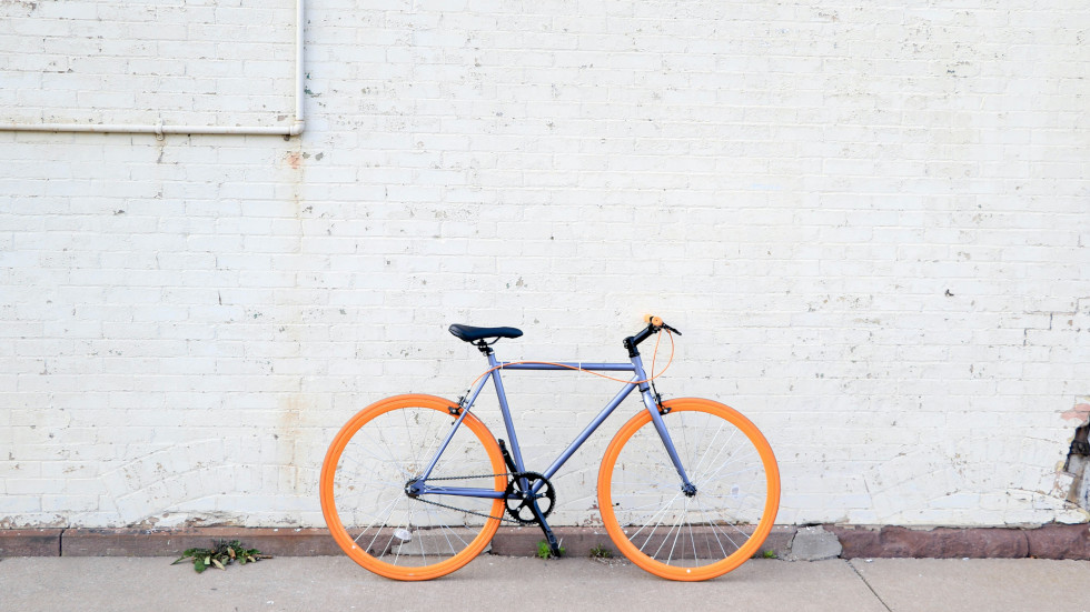 A blue framed single speed bicycle with orange wheels, against a brick wall painted white